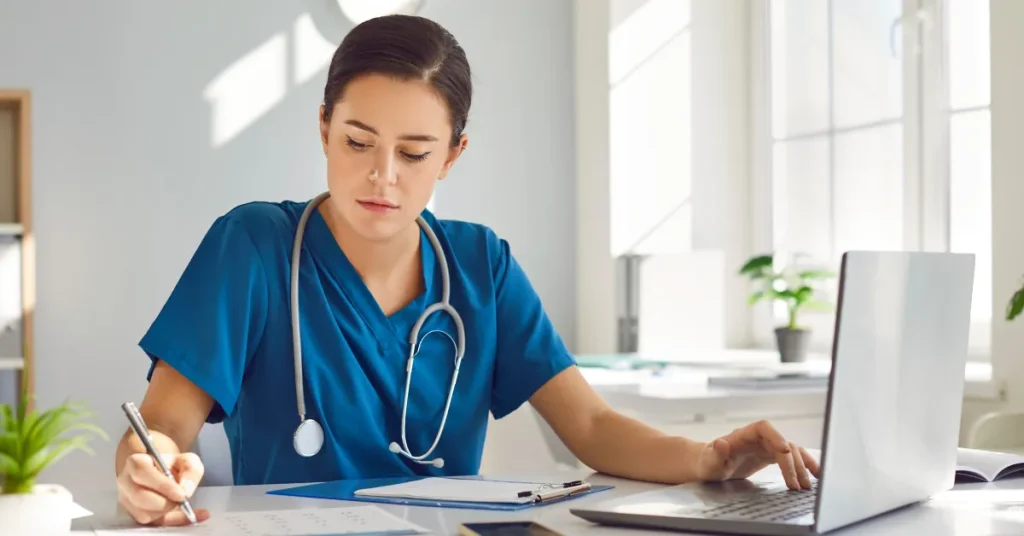 PA student in blue scrubs writes on a document while working on a laptop at an organized desk