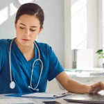 PA student in blue scrubs writes on a document while working on a laptop at an organized desk