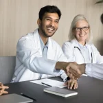 Young male PA shakes hands with his colleague across a conference table